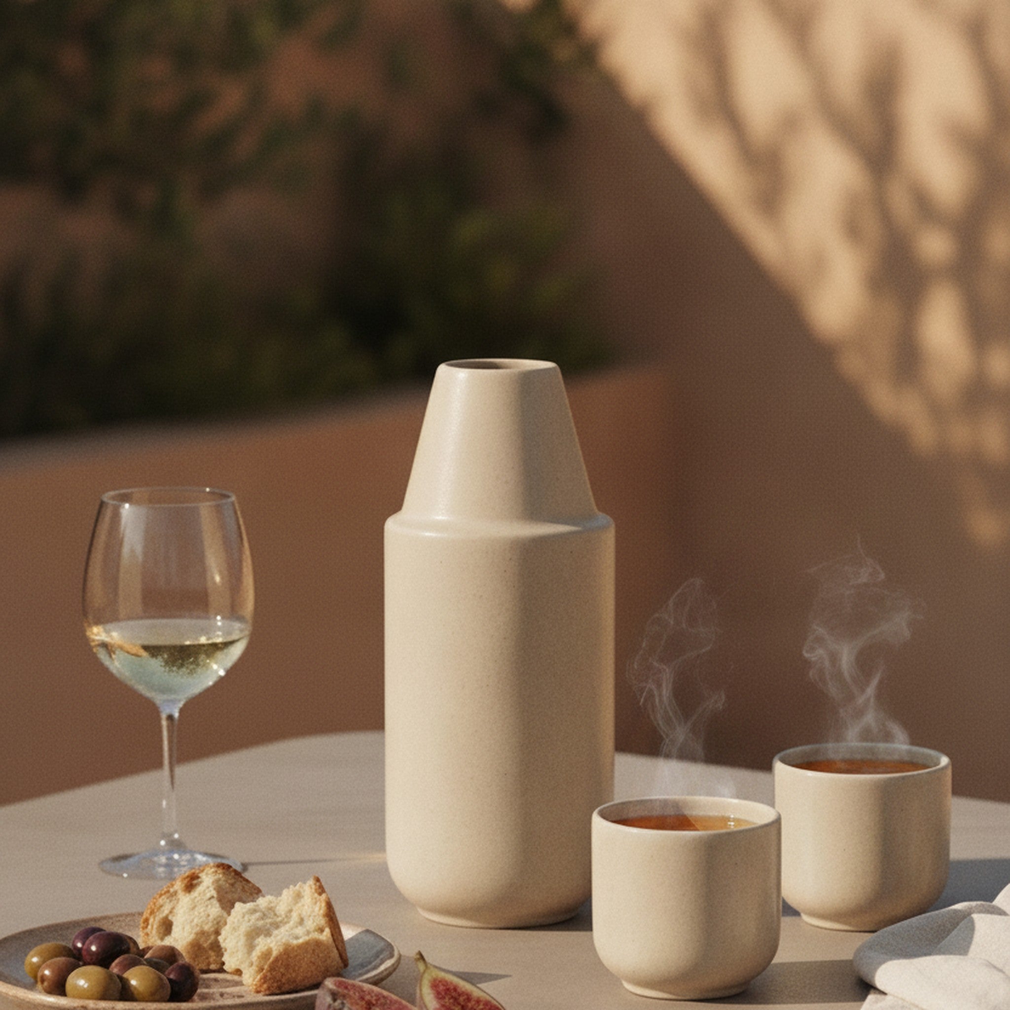 Handgegossene Keramik-Karaffe mit zwei Bechern auf einem mediterranen Tisch mit Weißwein, Brot und Oliven